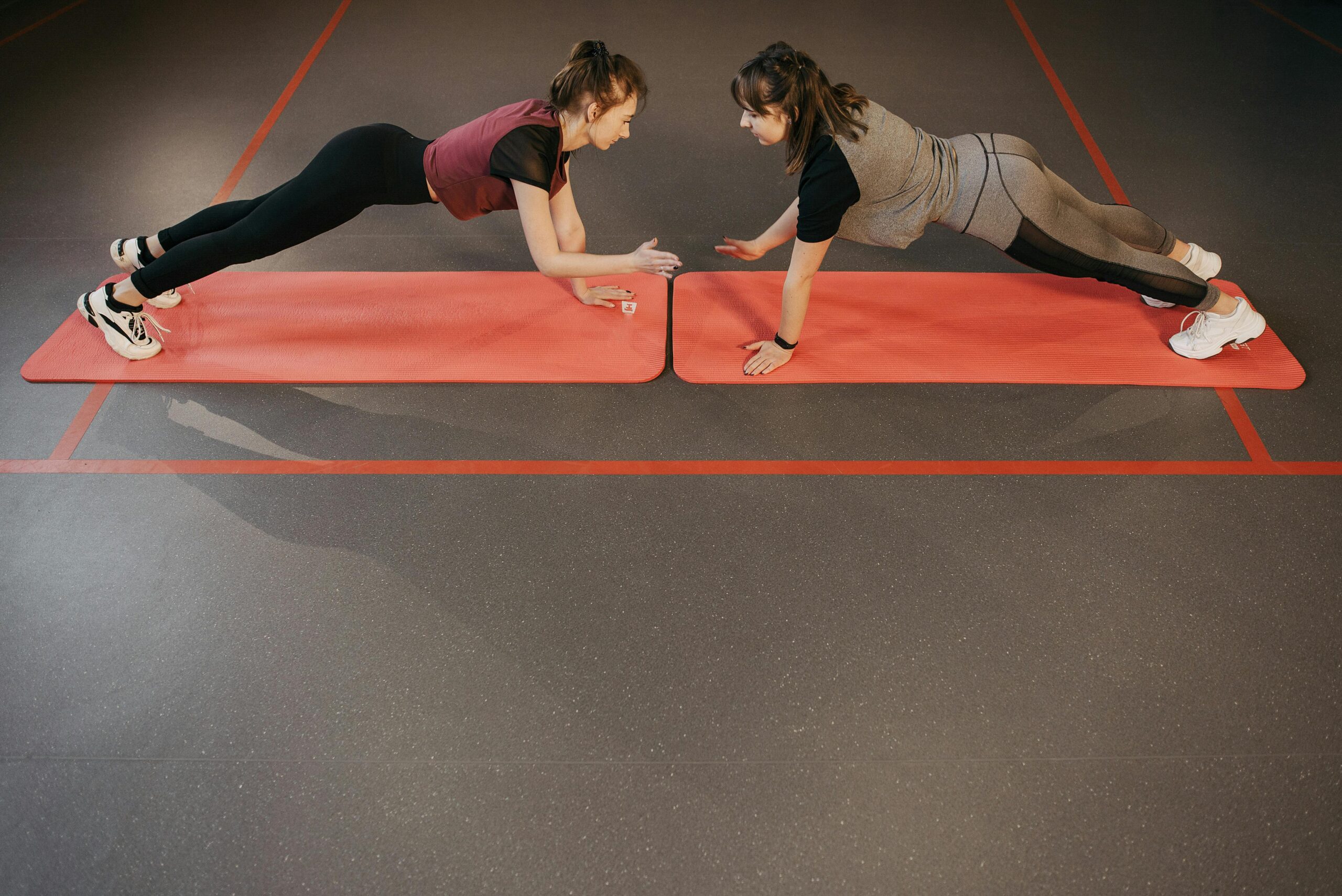 Two women planking on yoga mats in a gym, focusing on strength and teamwork.