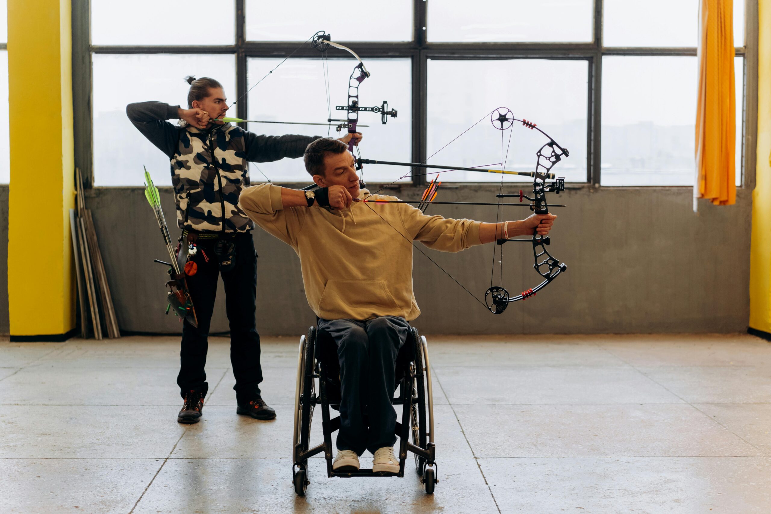 Two archers practice indoors, one using a wheelchair, showcasing determination and skill.